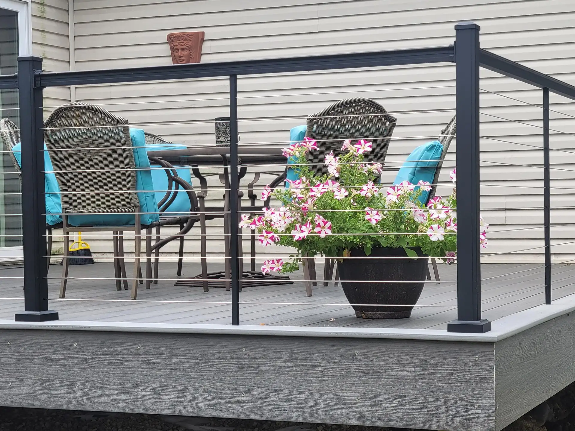 choosing composite decking colors Gray composite deck with black cable railing, wicker patio dining set with turquoise cushions and a planter of pink-and-white petunias.